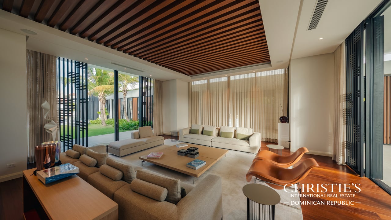 Furnished villa living room with garden-facing windows, a slatted wood ceiling, and sculptural orange lounge chairs.
