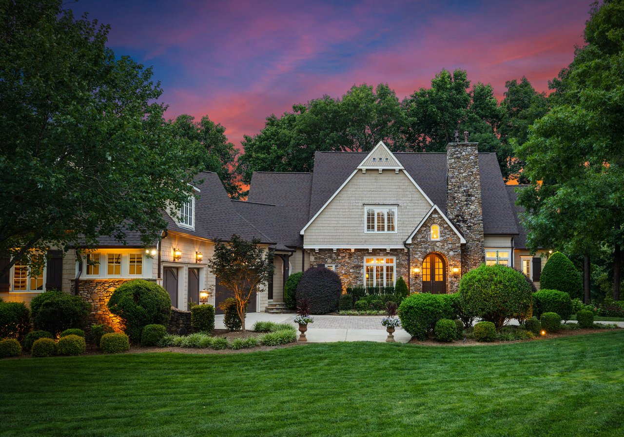Large stone-and-shingle home with a tall stone chimney and glowing windows at dusk.