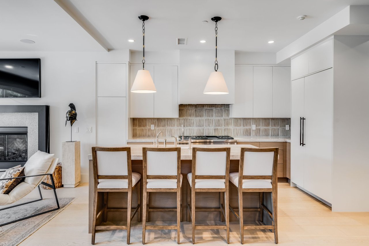 Modern kitchen with a large island, four wooden stools, white cabinets, and pendant lights.