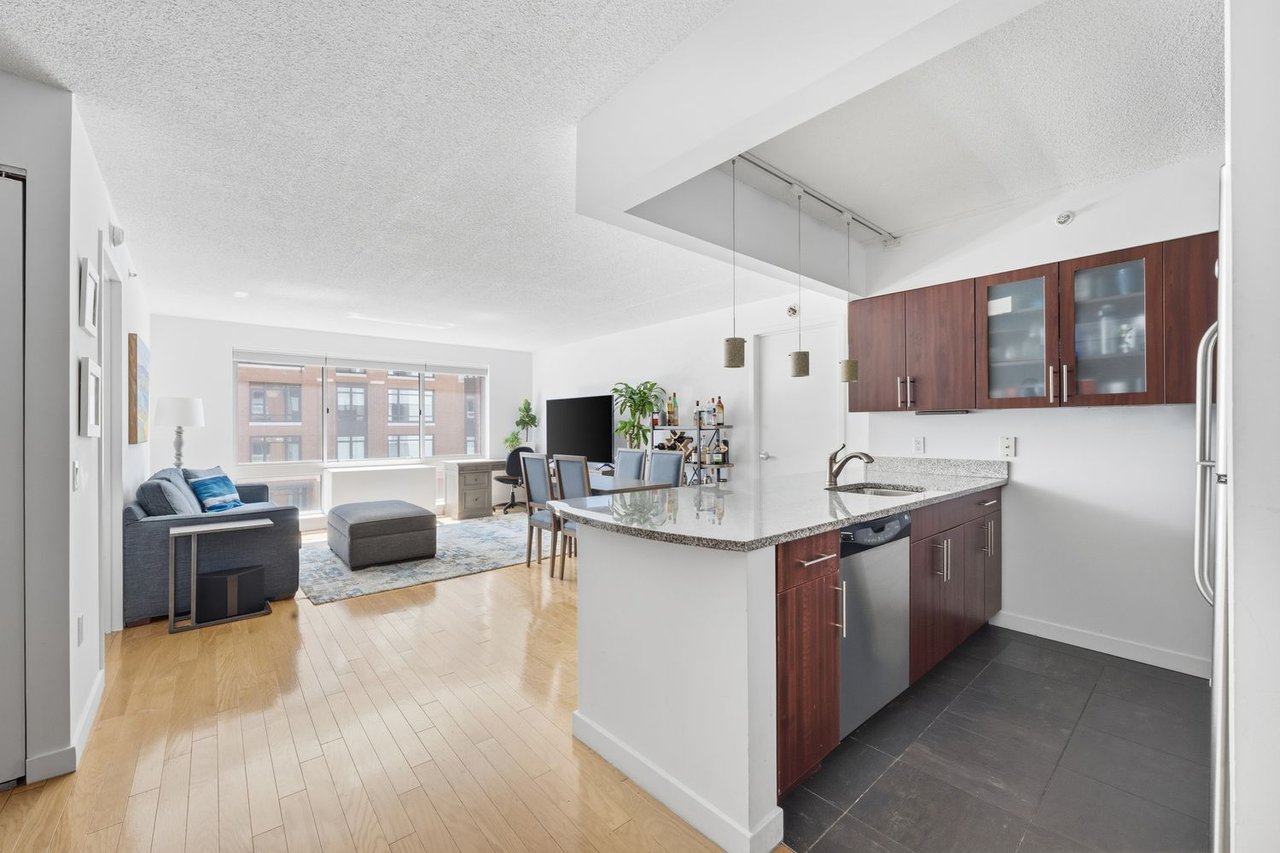 Open-plan condo kitchen and living area with a granite island, dark wood cabinets, and large windows.