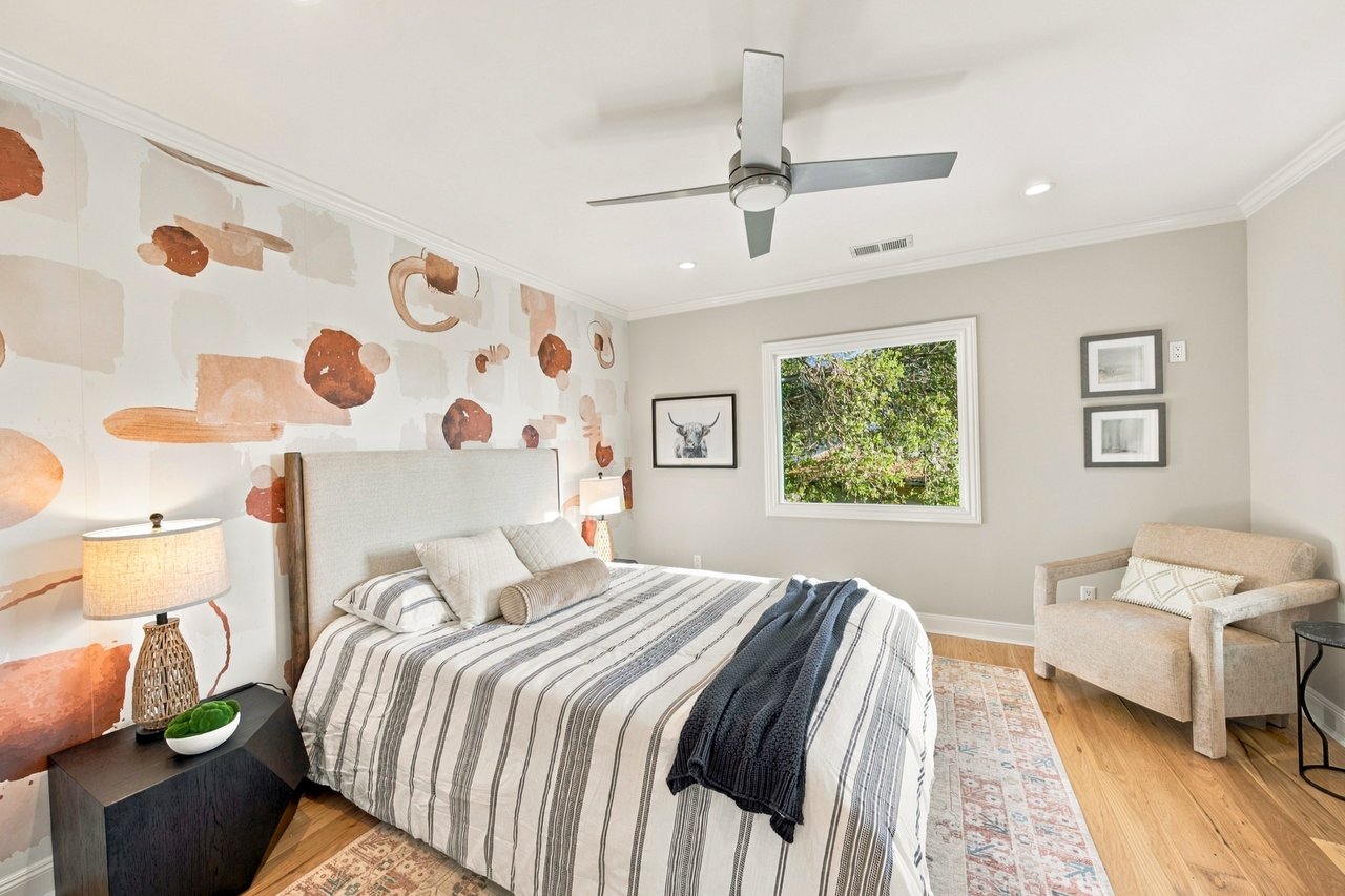 Staged bedroom with striped bedding, patterned accent wallpaper, and an armchair beside a window.