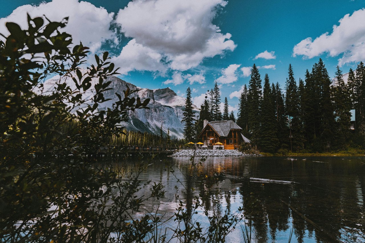 Wooden lodge beside a mountain lake, with evergreen trees and a partly snow-covered mountain beyond.