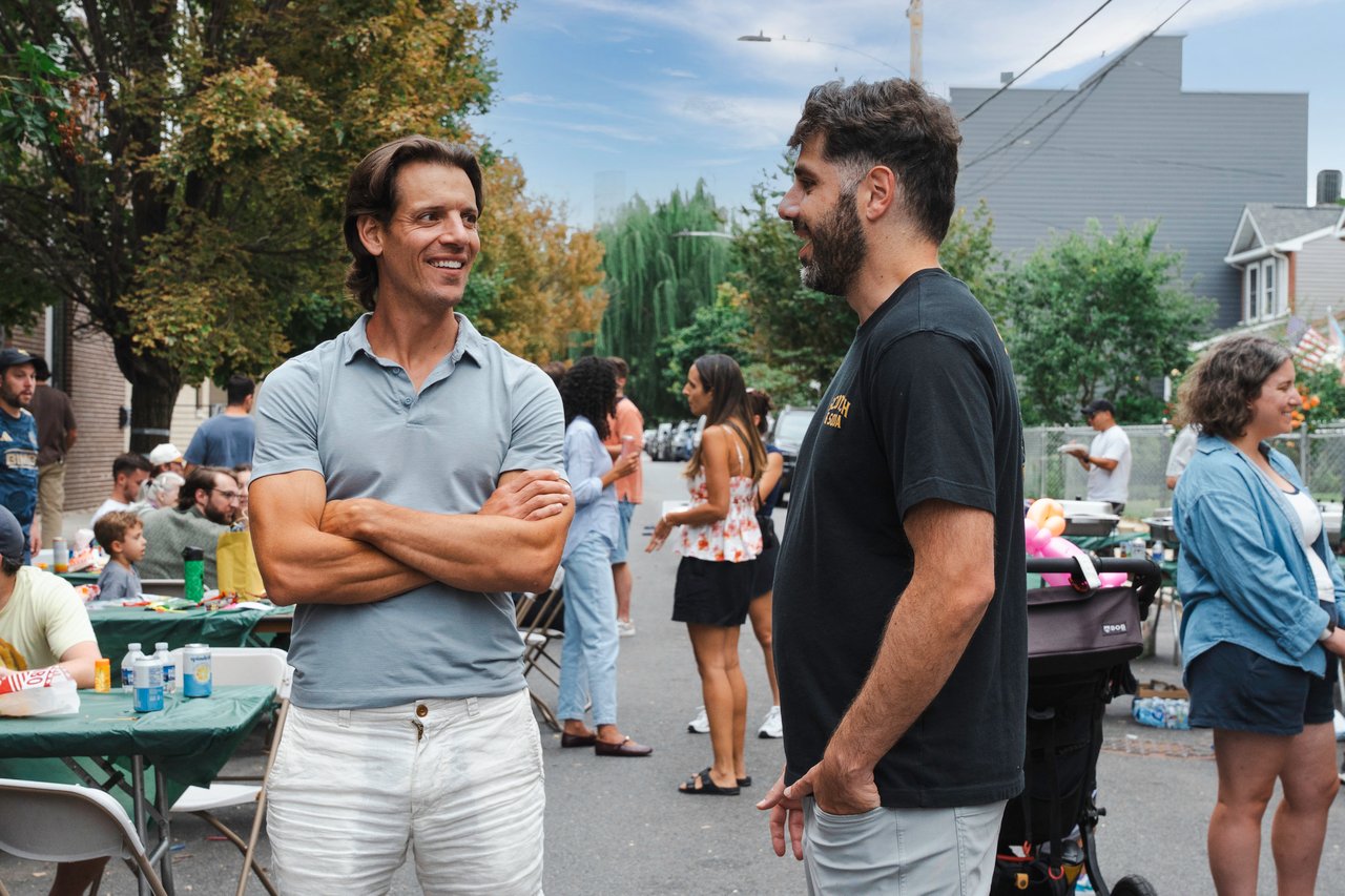 Neighbors chat at a block party on a residential street, with families and picnic tables behind them.