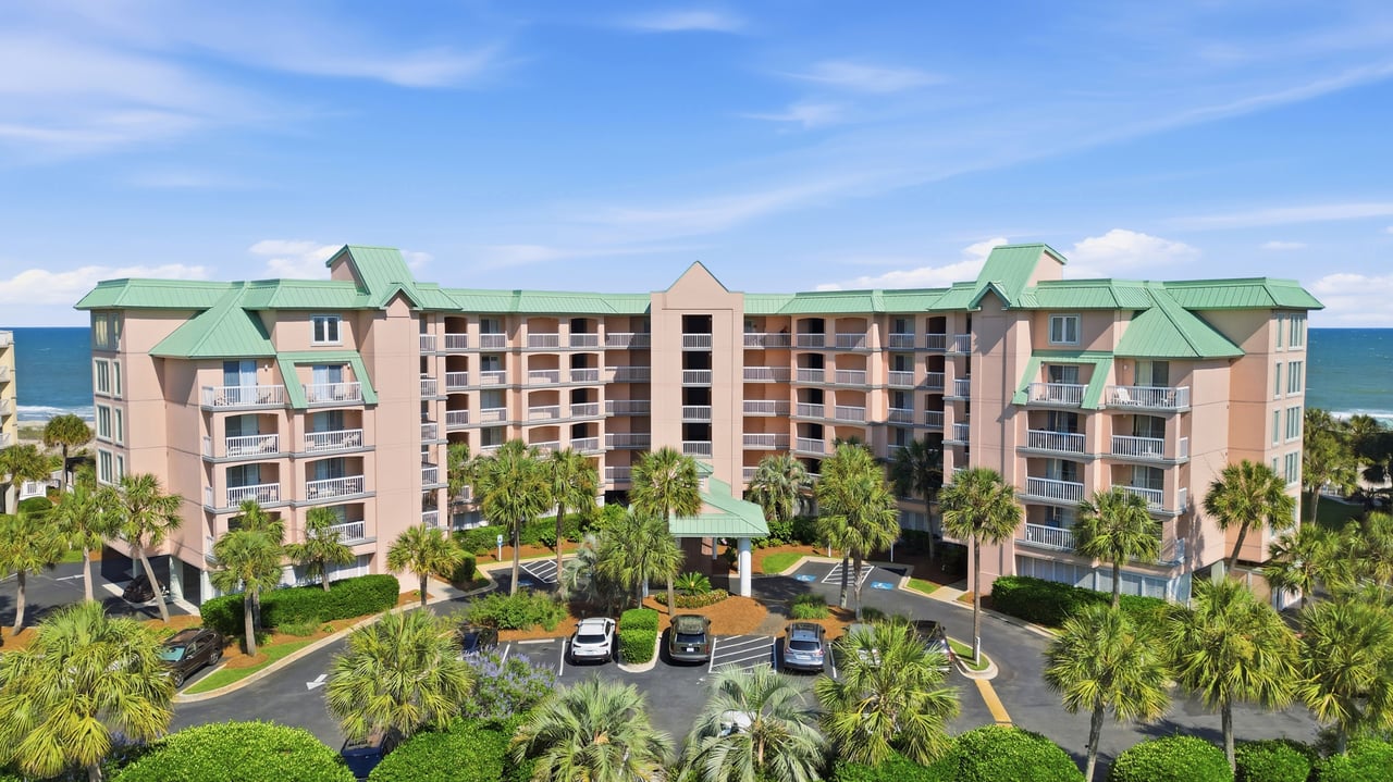 Coastal condominium with green roofs and balconies, palm trees in front and ocean behind.