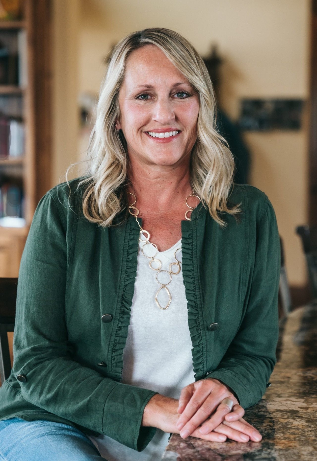 Shelly Albers smiling in a dark green jacket and white top, seated at a counter.