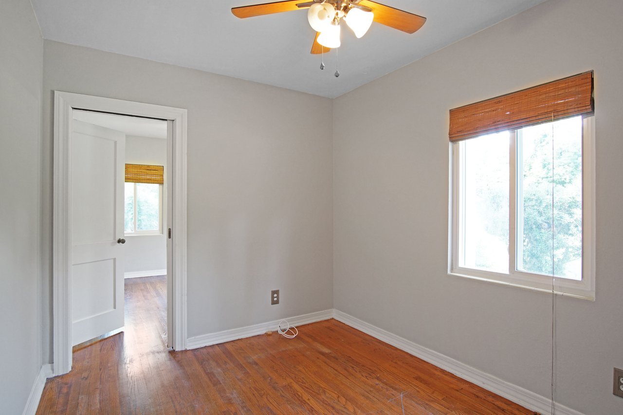 Empty room with light gray walls and wooden floor, featuring a ceiling fan and window with bamboo blinds. Doorway leads to another bright room.