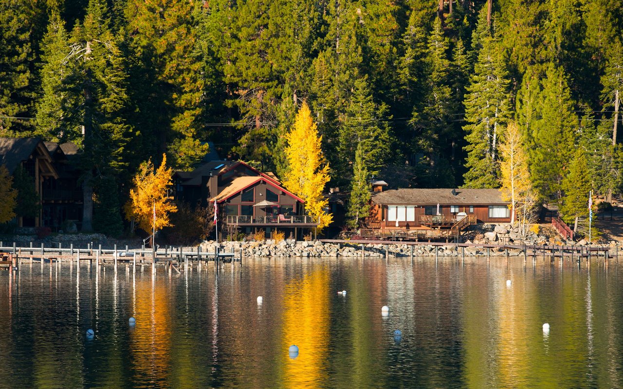 Lakeside homes and docks reflected in calm water, backed by evergreen forest and golden autumn trees.