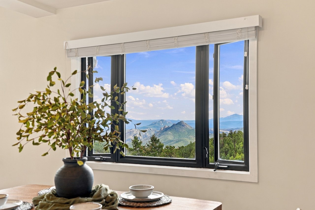 Black-framed window overlooking forested mountains, with a potted plant and table in the foreground.