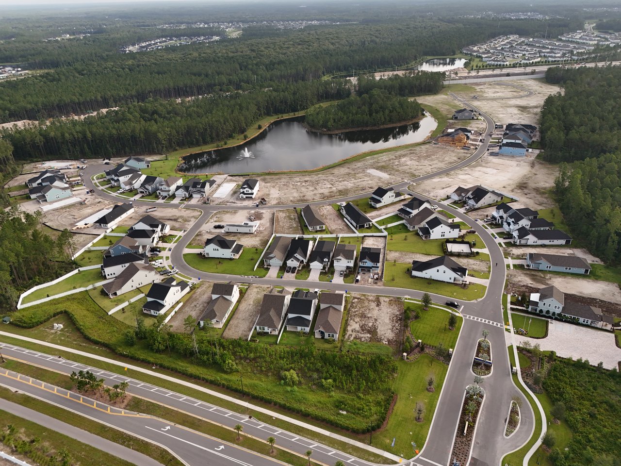 Aerial view of new homes surrounding a pond with a fountain, with wooded land beyond.