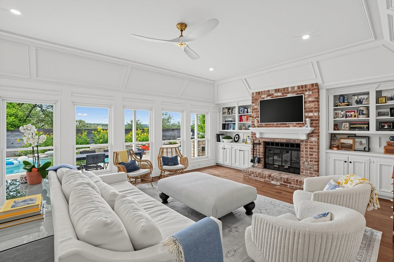 Living room with a brick fireplace, built-in bookshelves, and tall windows overlooking the pool.
