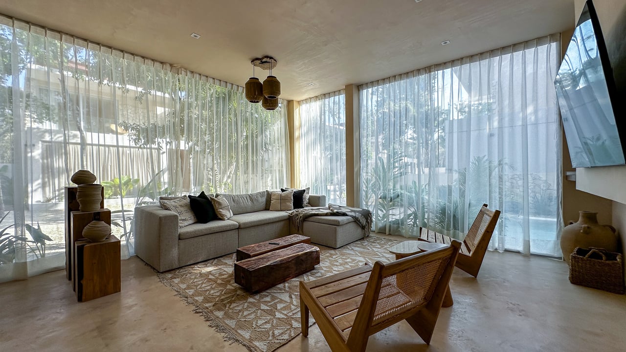 Living room with a beige sectional, woven chairs, and sheer curtains overlooking tropical greenery.