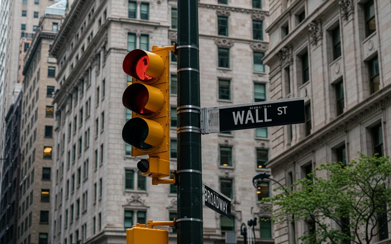 Wall Street and Broadway street signs beside a red traffic light and tall Manhattan buildings.