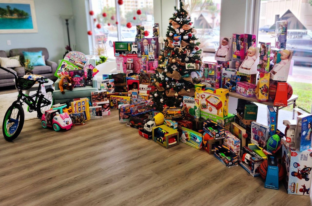 Christmas tree surrounded by donated toys, dolls, games, and bicycles in a bright living room.