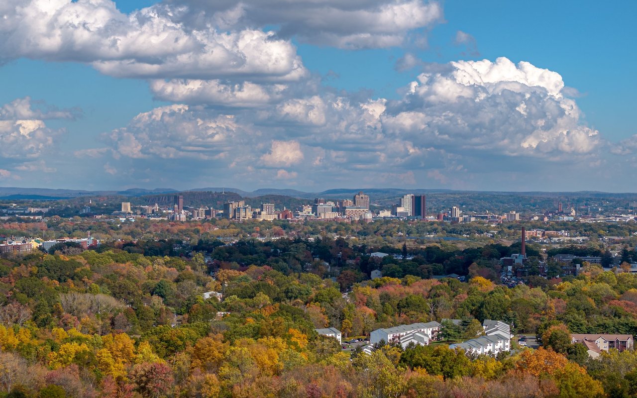 New Haven skyline beyond colorful autumn trees beneath a cloud-filled sky.