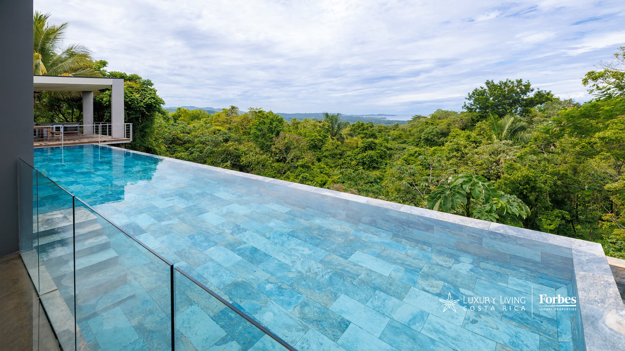 Infinity pool with glass railings overlooking lush tropical forest and distant hills.