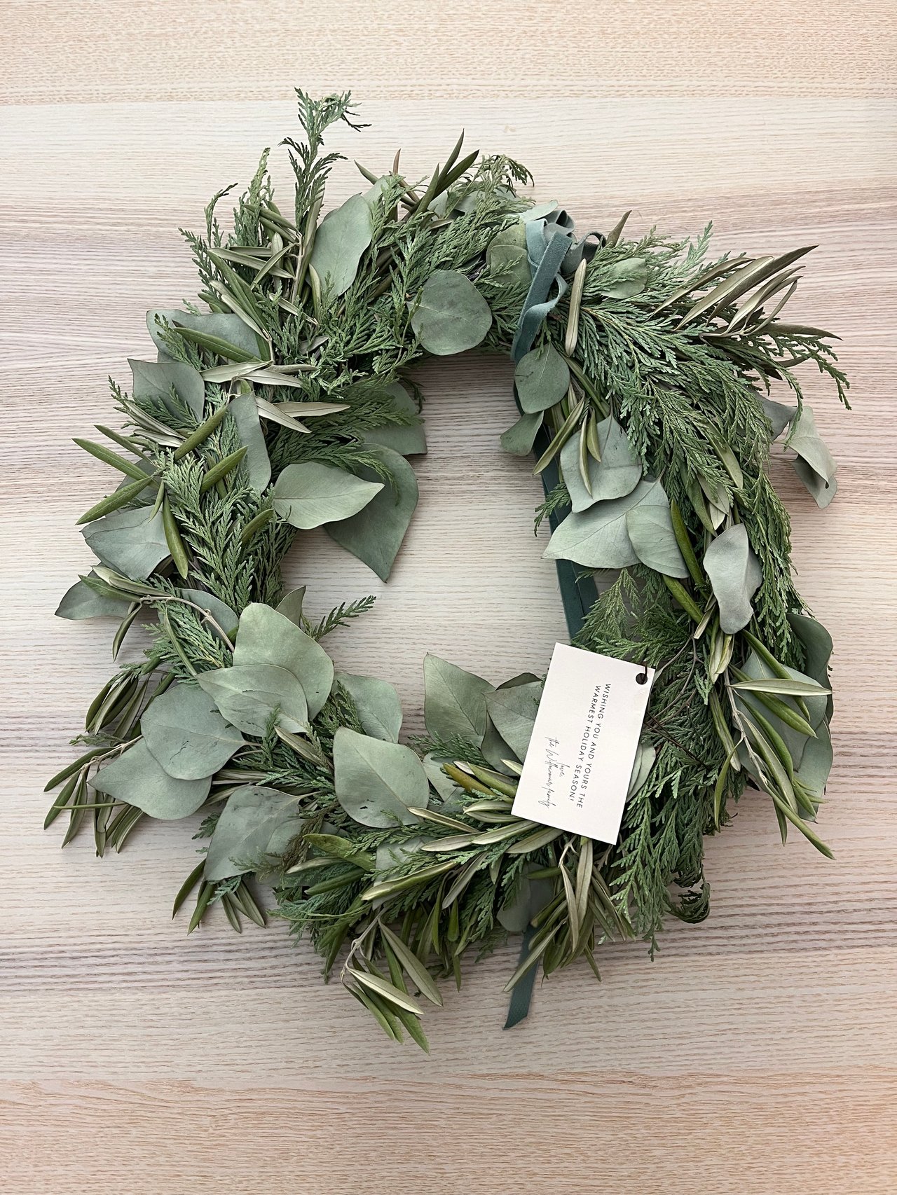 Evergreen holiday wreath with eucalyptus leaves, teal ribbon, and a seasonal greeting tag.