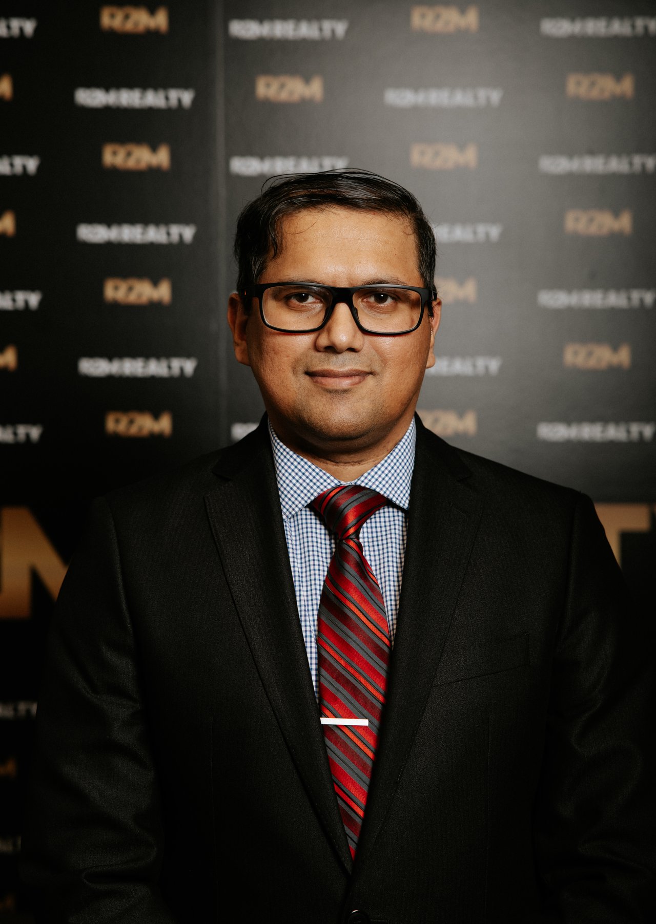 Azizur Rasel in glasses and a black suit with a red striped tie against an RPM Realty backdrop.