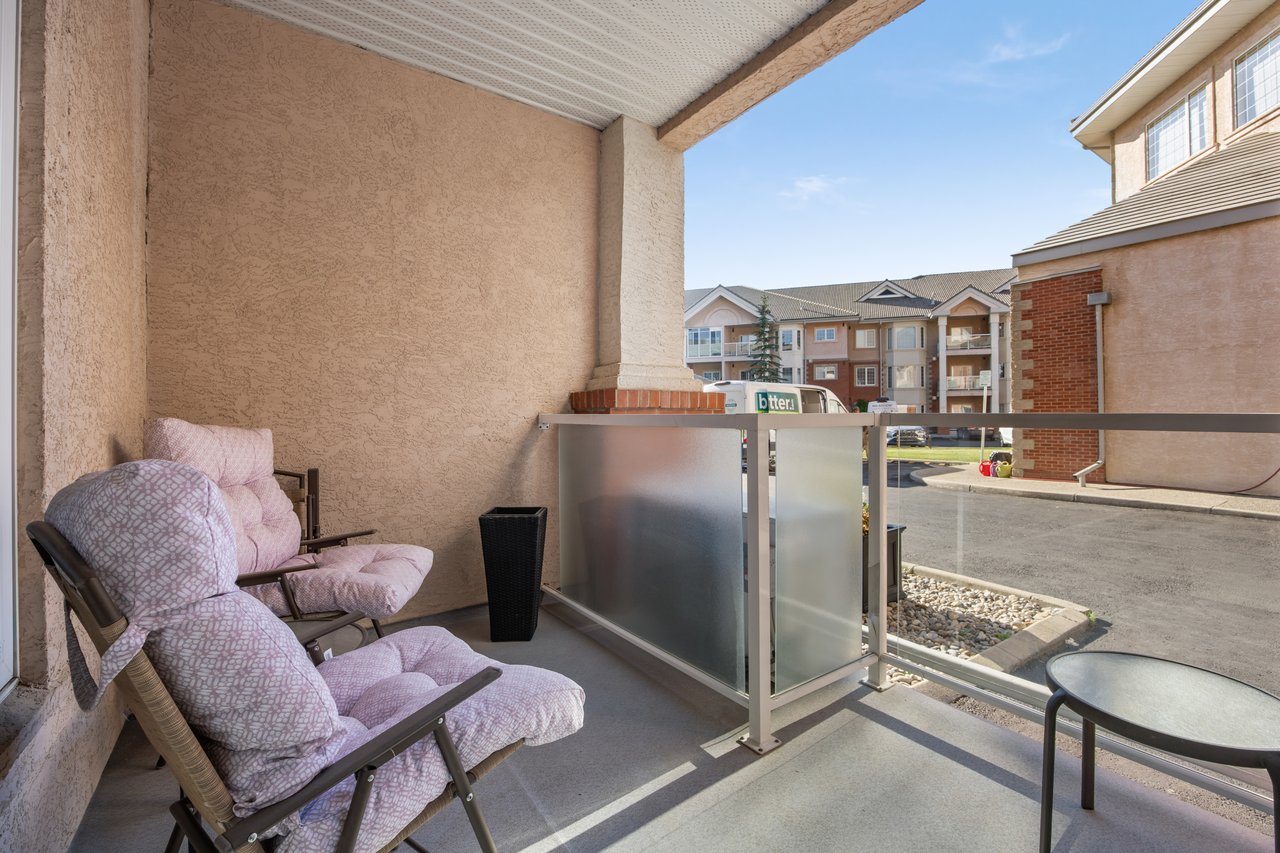 Covered apartment balcony with cushioned lounge chairs and a frosted-glass railing overlooking neighboring buildings.