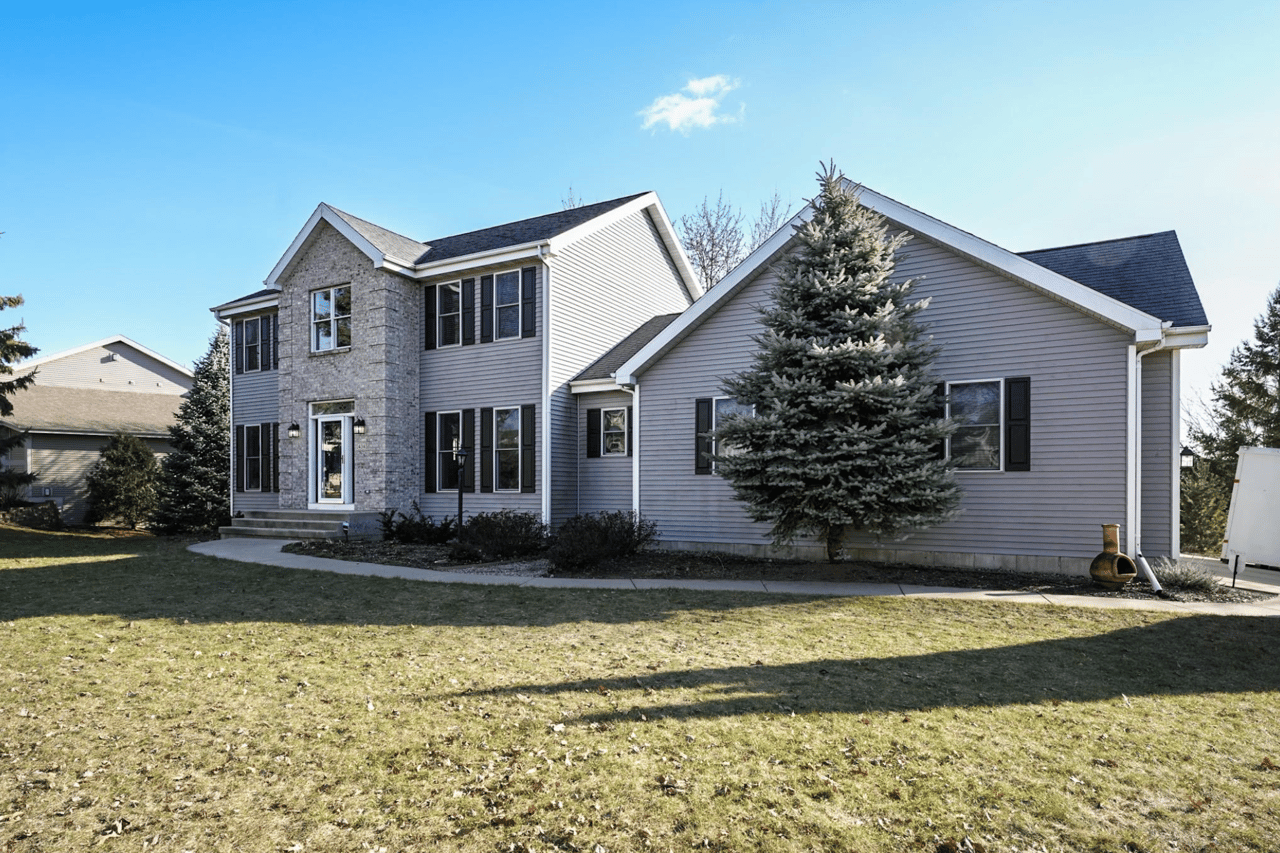 Two-story gray-sided home with a stone entrance facade and a large spruce tree in the yard.
