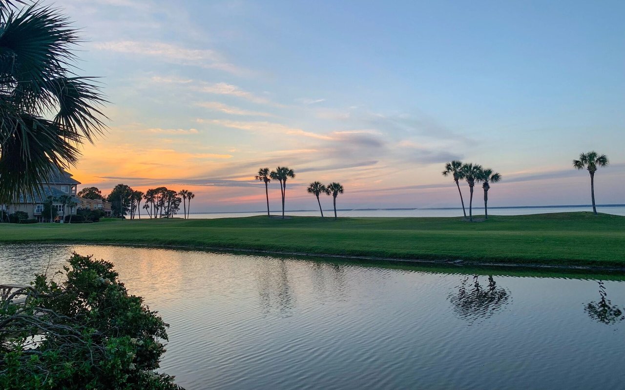 Coastal golf course at sunset, with a pond, palm-lined fairway, and ocean horizon.