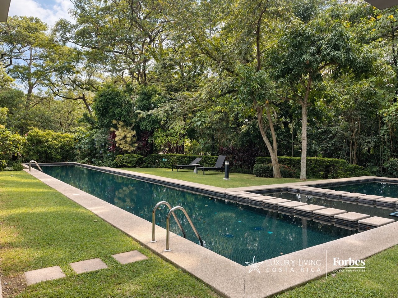 Swimming pool in a lush tropical garden, with submerged stepping stones and two loungers.