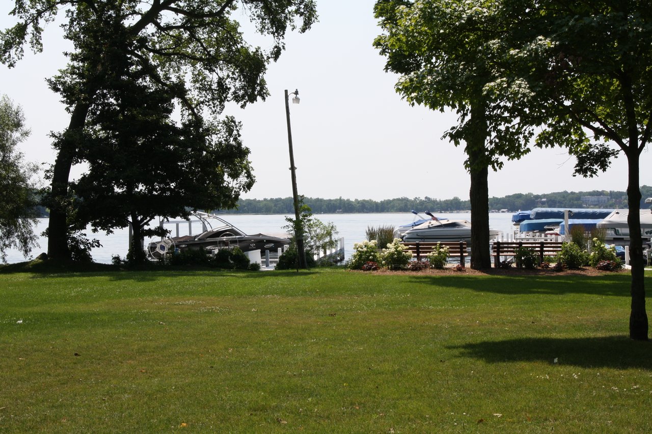 Lakeside lawn with mature trees, flowering shrubs, docks, and boats along the water.