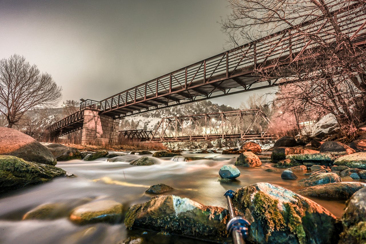 Rocky river flowing beneath two pedestrian bridges, with leafless trees and snow-dusted hills.