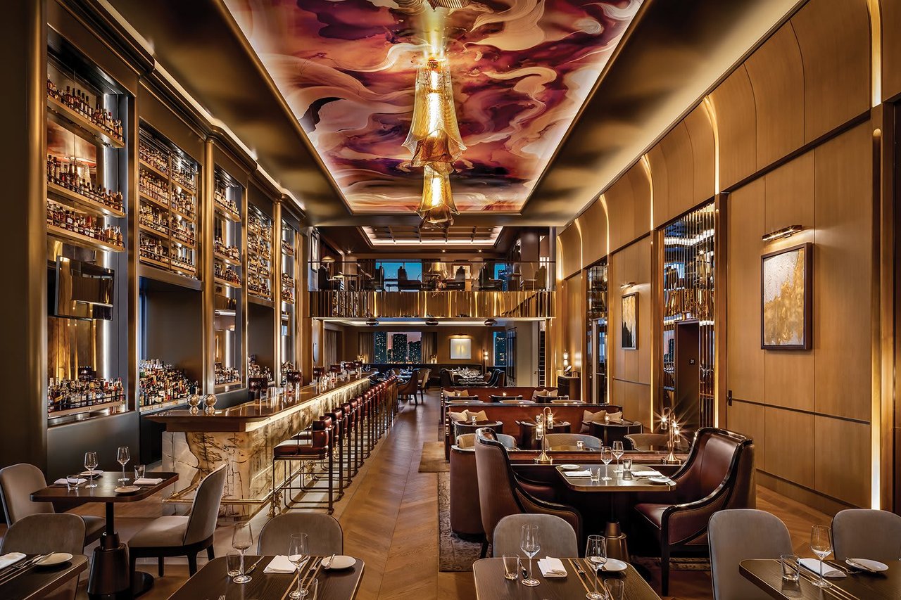 Upscale restaurant dining room with a bottle-lined bar and colorful ceiling artwork.