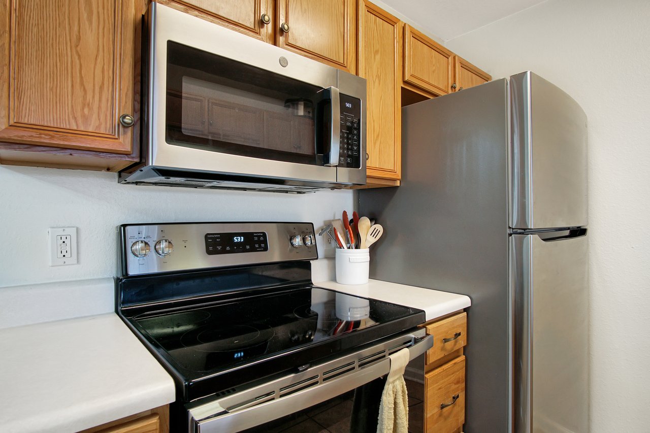 Modern kitchen corner featuring stainless steel appliances, wooden cabinets, and white countertops. A jar holds utensils beside the stove. Cozy and tidy.