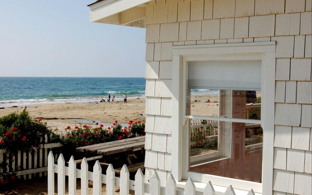 White shingle beach house overlooking the ocean, with a picket fence and flowering shrubs.