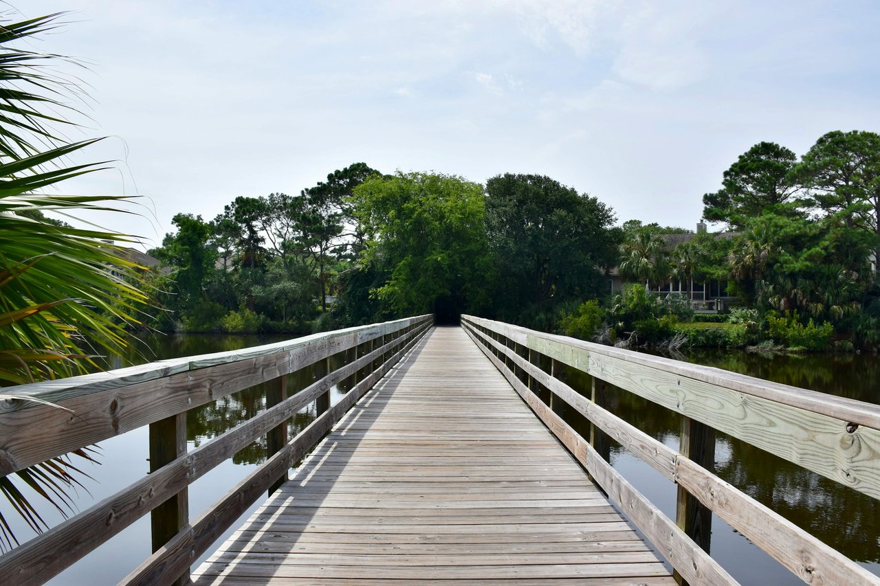 Wooden boardwalk crosses calm water toward a tree-lined trail.