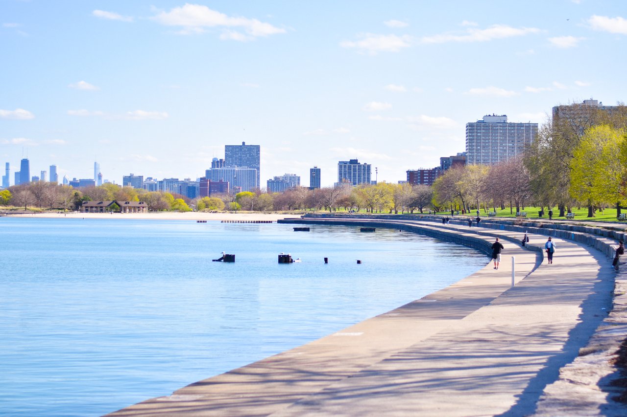 Lakefront promenade curves beside blue water toward the Chicago skyline, lined with spring trees.