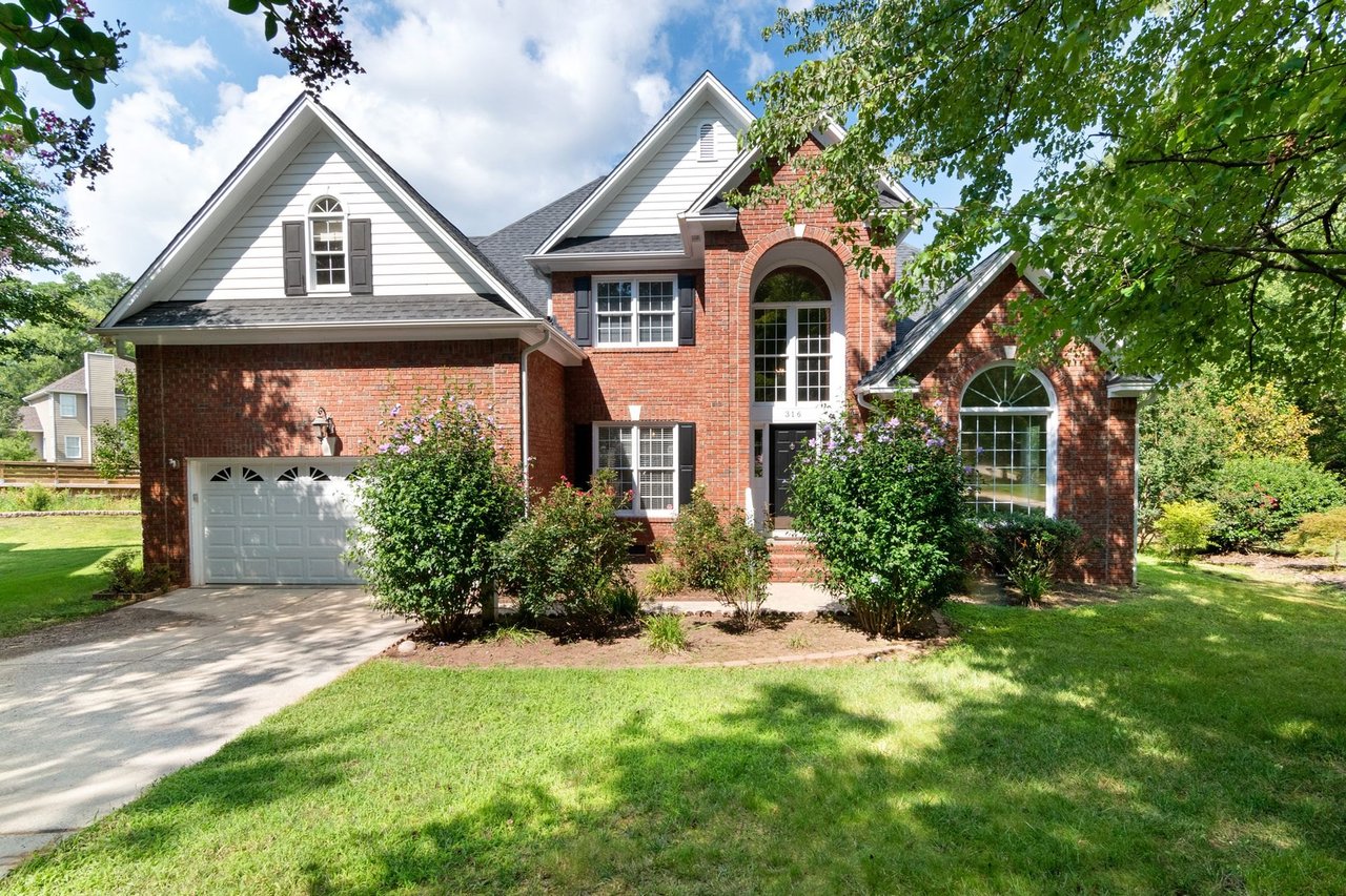 Two-story red brick house with a two-car garage, front shrubs and a green lawn under a blue sky with clouds