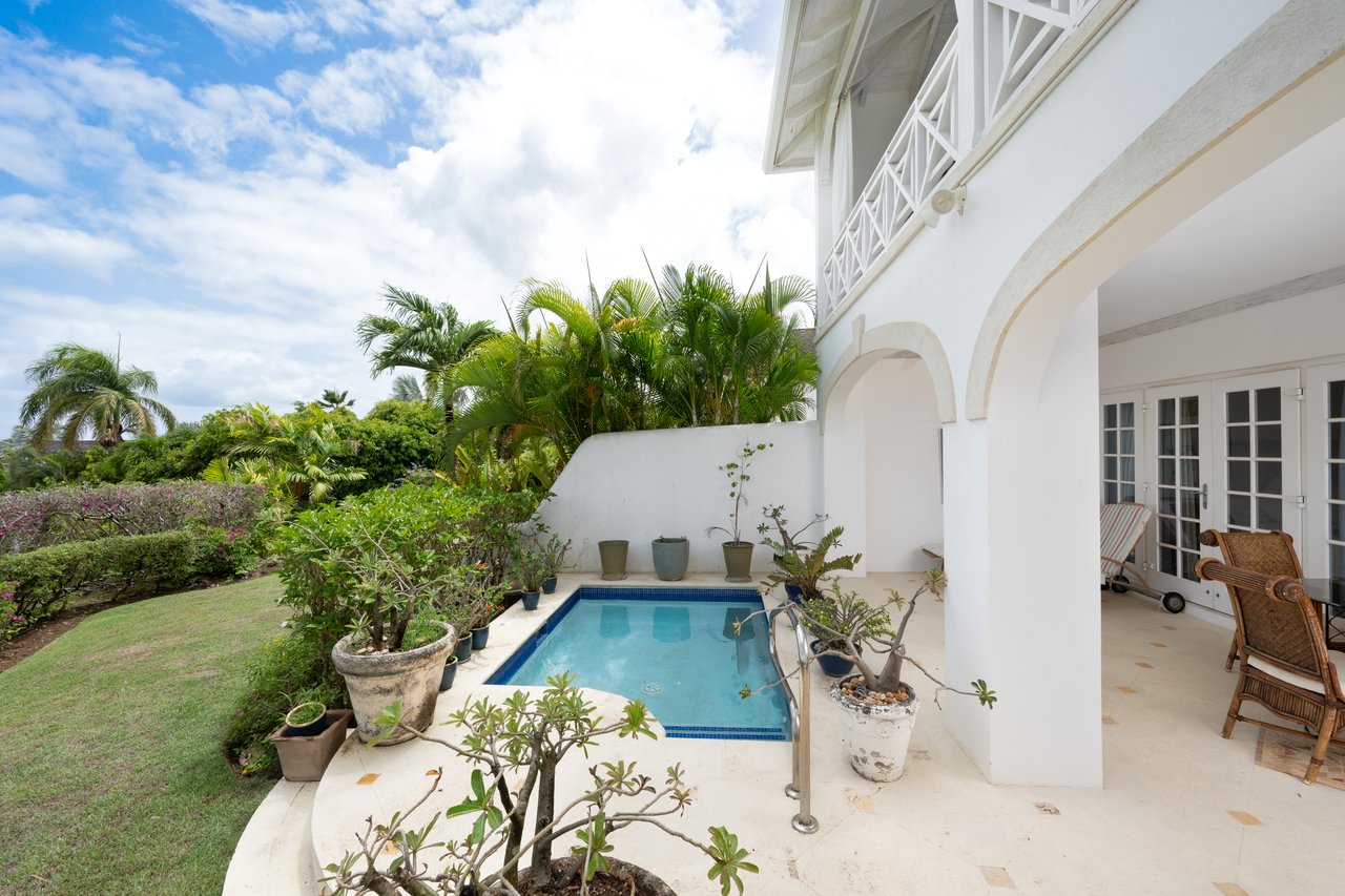 Villa pool beside a white arched patio, with wicker chairs, potted plants and tropical garden.