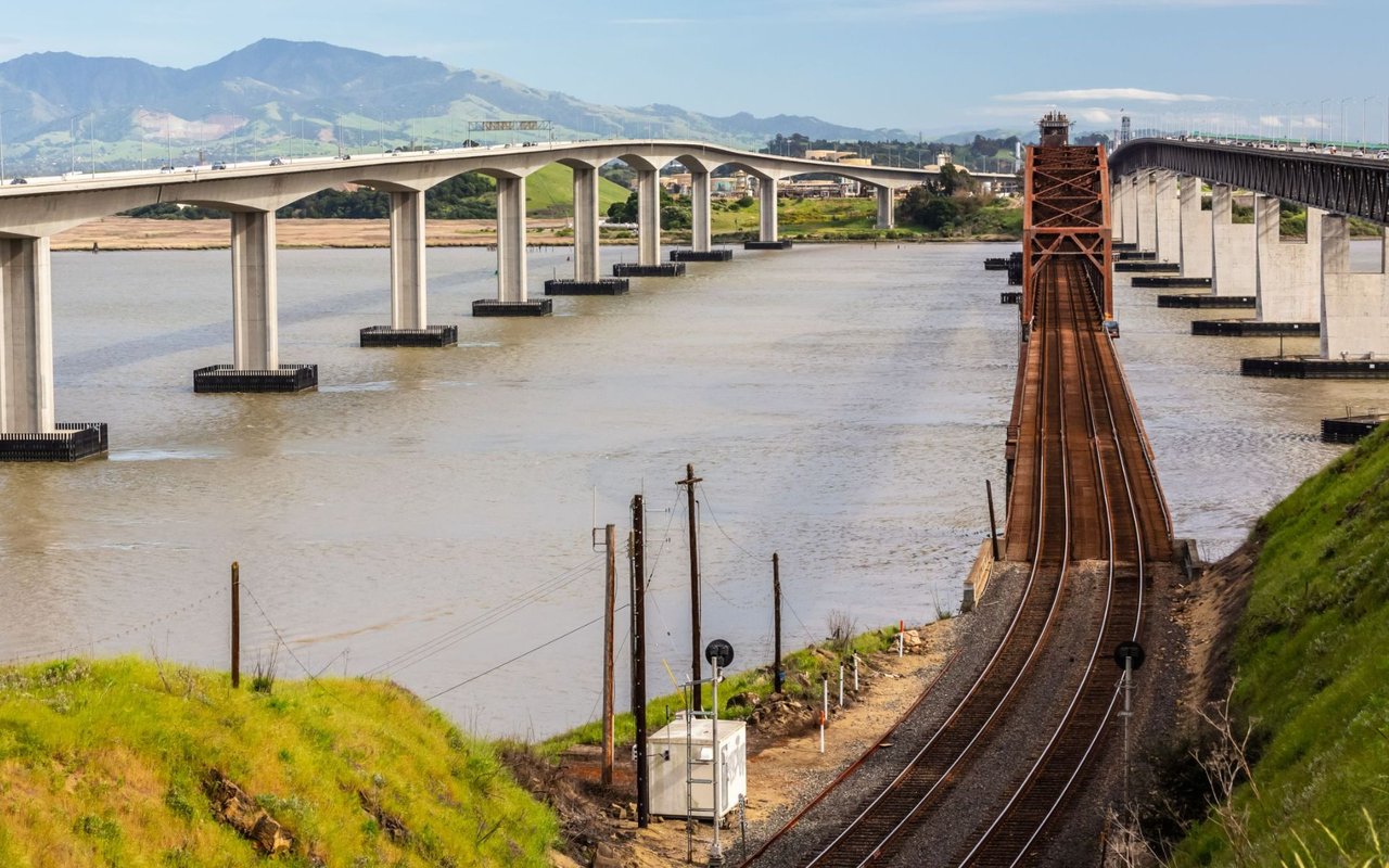 Bridges cross the water near Benicia, with railway tracks in the foreground and green hills beyond.