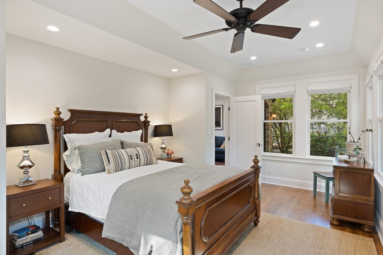 Bedroom with a dark wood four-poster bed, gray bedding, hardwood floors, and large windows overlooking greenery.