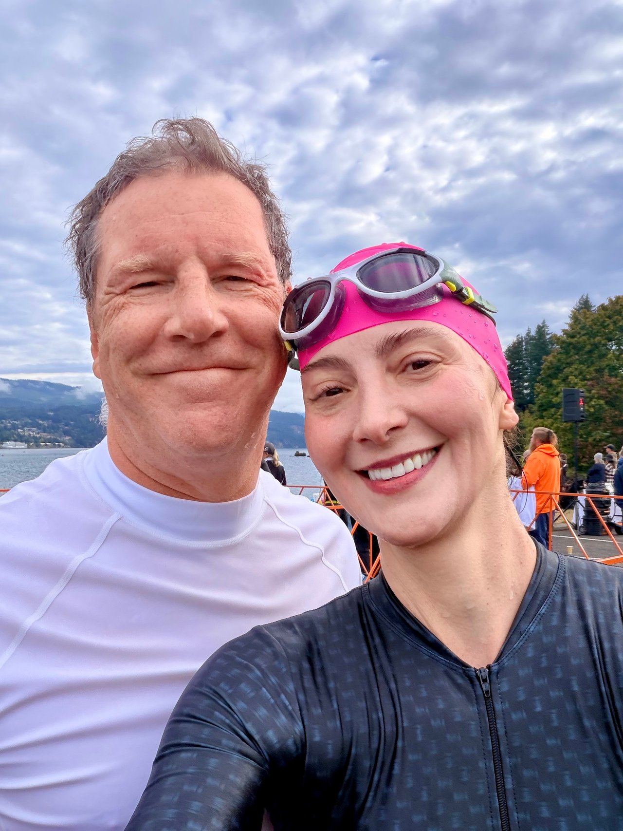 Two adults smile for a selfie by the water; one wears a pink swim cap and goggles.