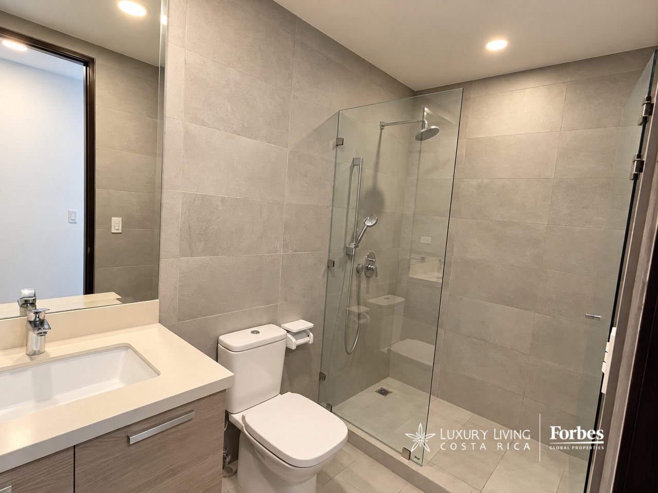 Modern tiled bathroom with a glass walk-in shower, rainfall showerhead, vanity, and toilet.