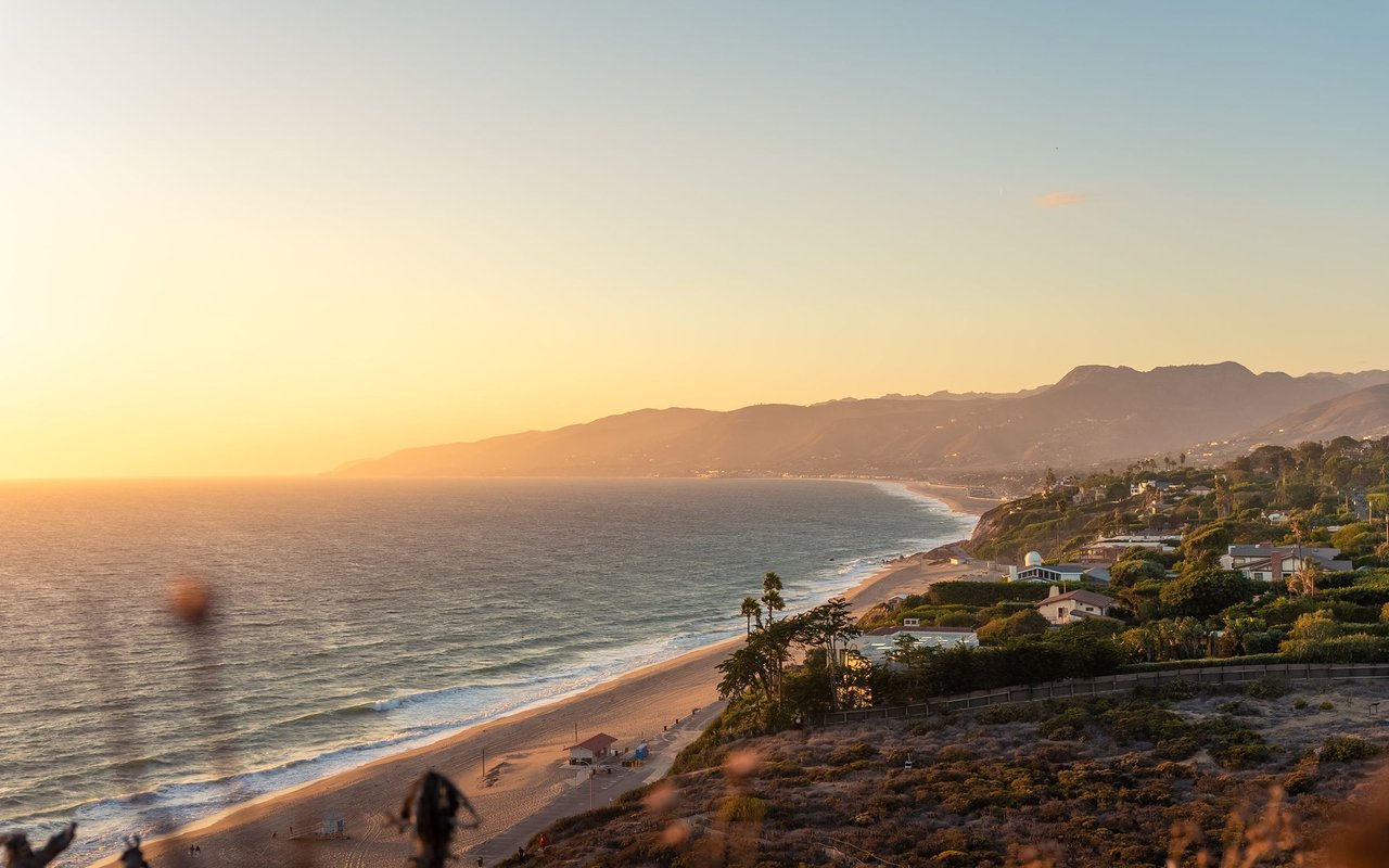 Malibu coastline at sunset, with a sandy beach, oceanfront homes, and distant mountains.