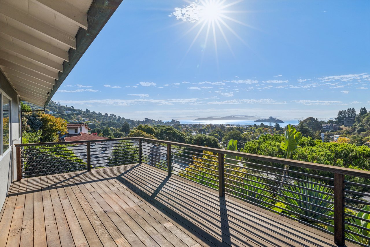 Wooden deck overlooking a green hillside neighborhood and bay, with distant islands and a bridge.