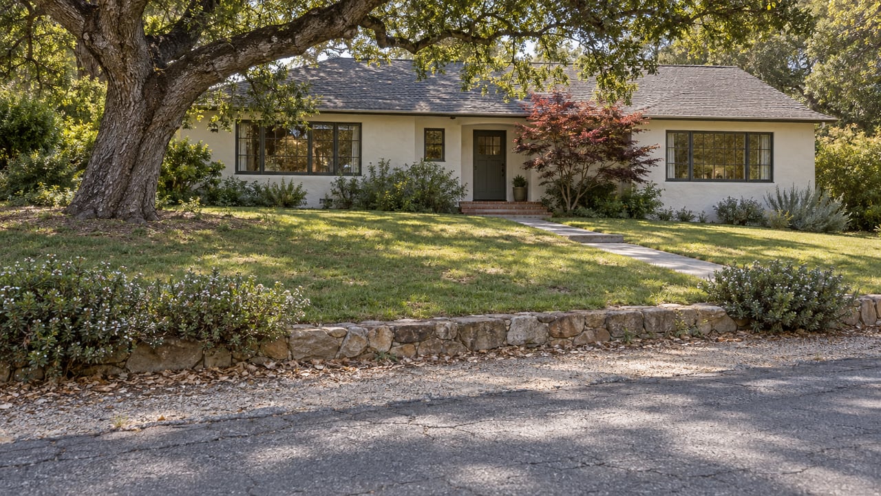 Single-story stucco ranch beyond a rising lawn, with an oak canopy overhead and a short concrete walk.
