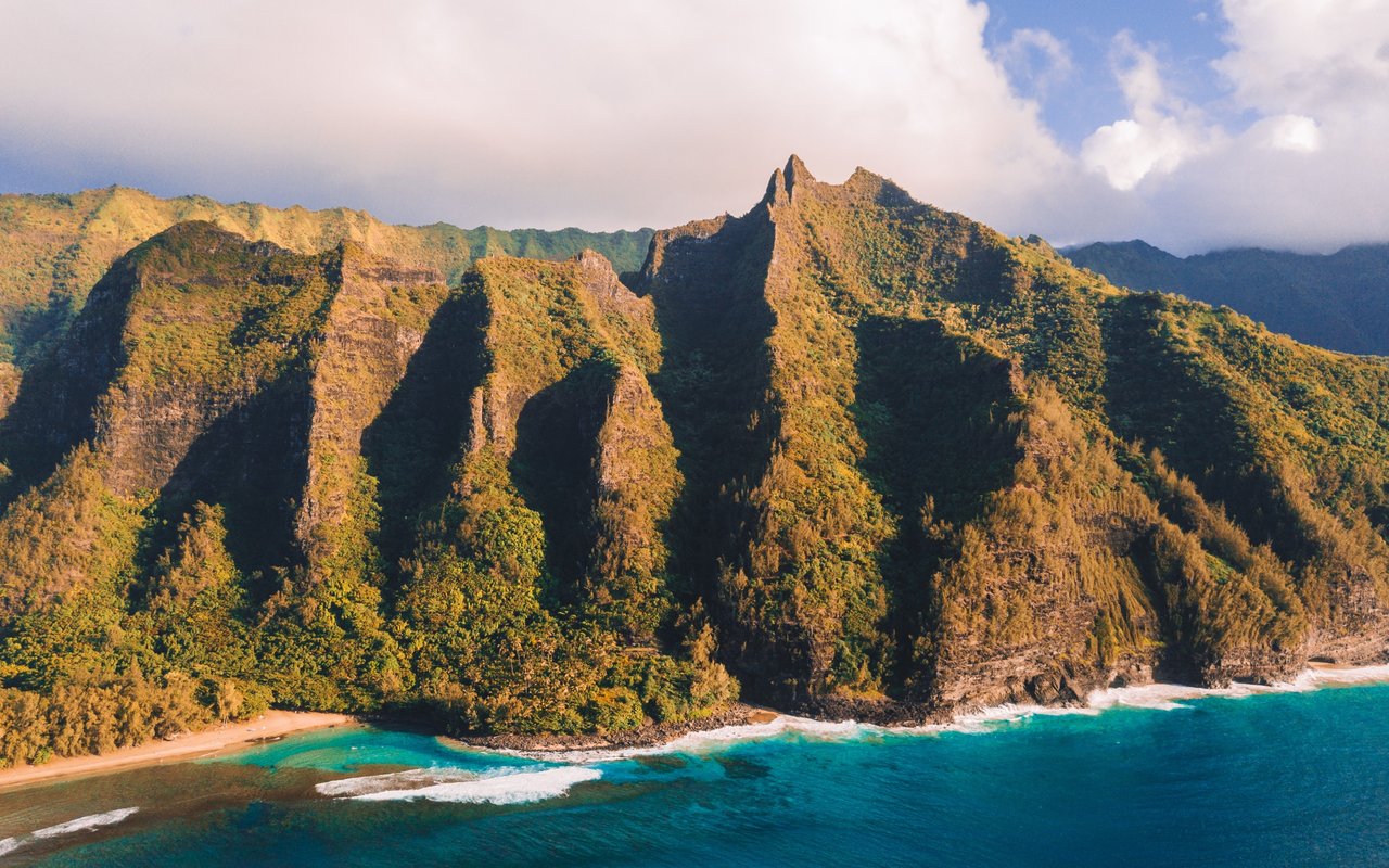 Aerial view of rugged green coastal cliffs above turquoise water and narrow beaches.