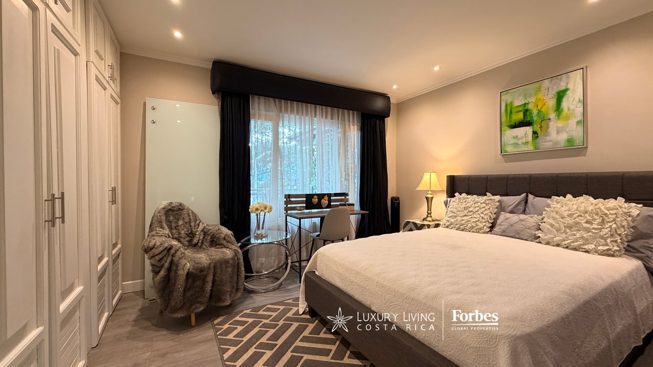 Bedroom with upholstered bed, built-in white wardrobes, and desk beside a curtained window.