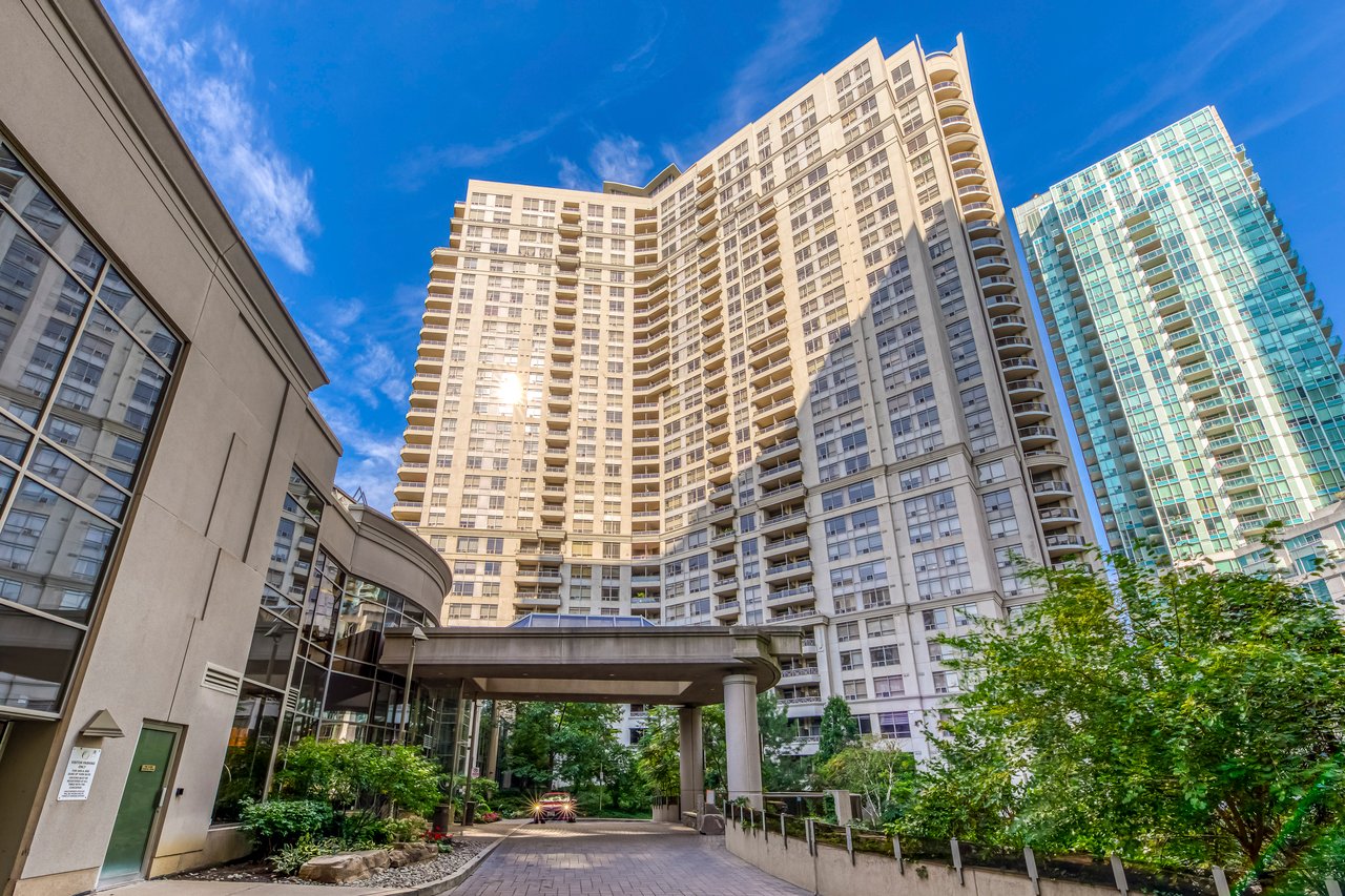 High-rise condominiums viewed from a landscaped driveway beneath a bright blue sky.