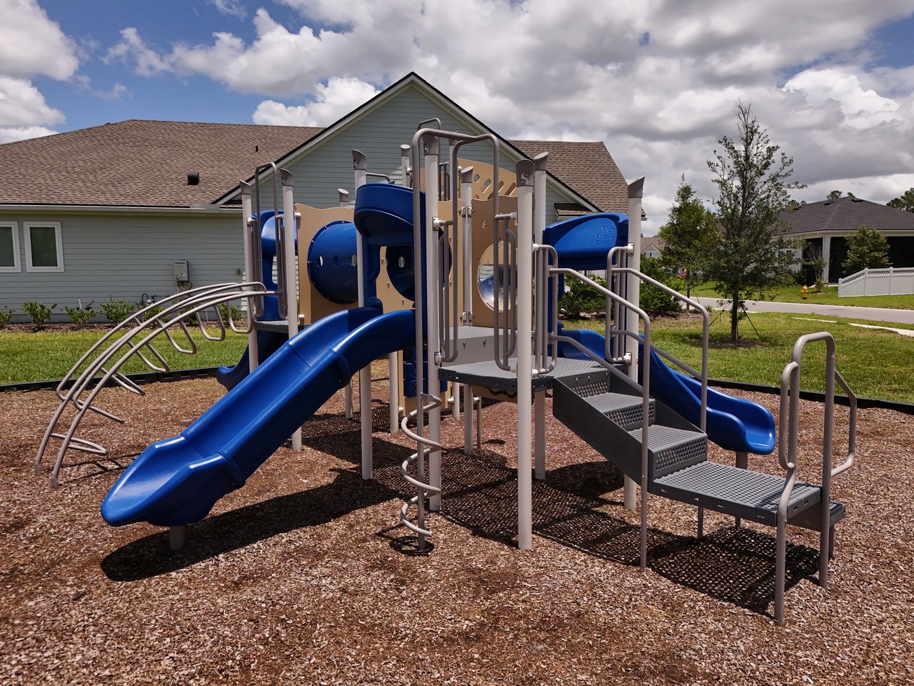 Community playground with blue slides and climbing equipment beside neighborhood homes.