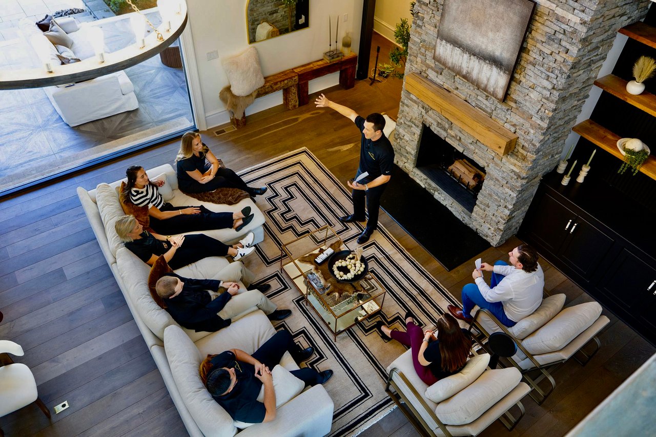 A real estate agent presents to a seated group around white sofas in a living room with a stone fireplace.