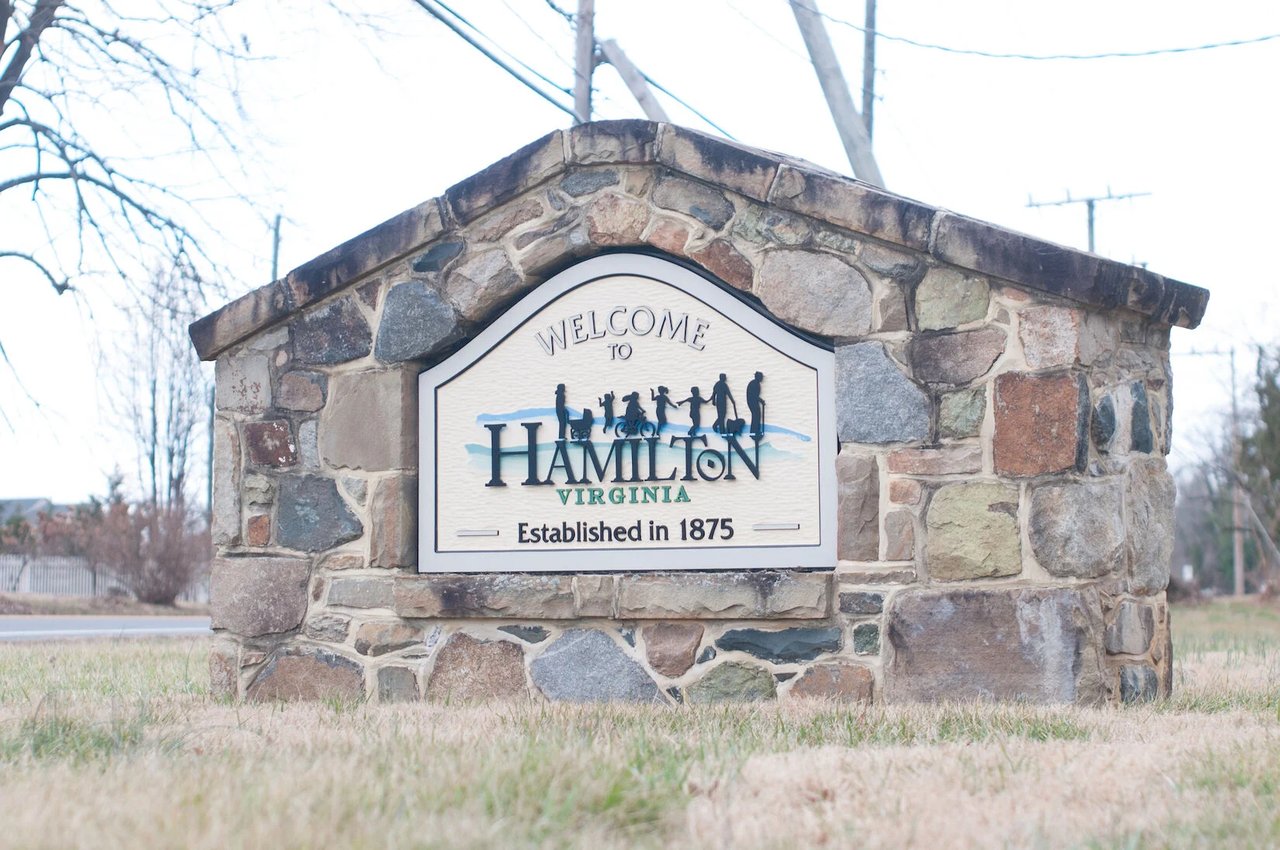 Stone "Welcome to Hamilton, Virginia — Established in 1875" monument sign with a silhouette of walkers, cyclists and families along a roadside in Loudoun County