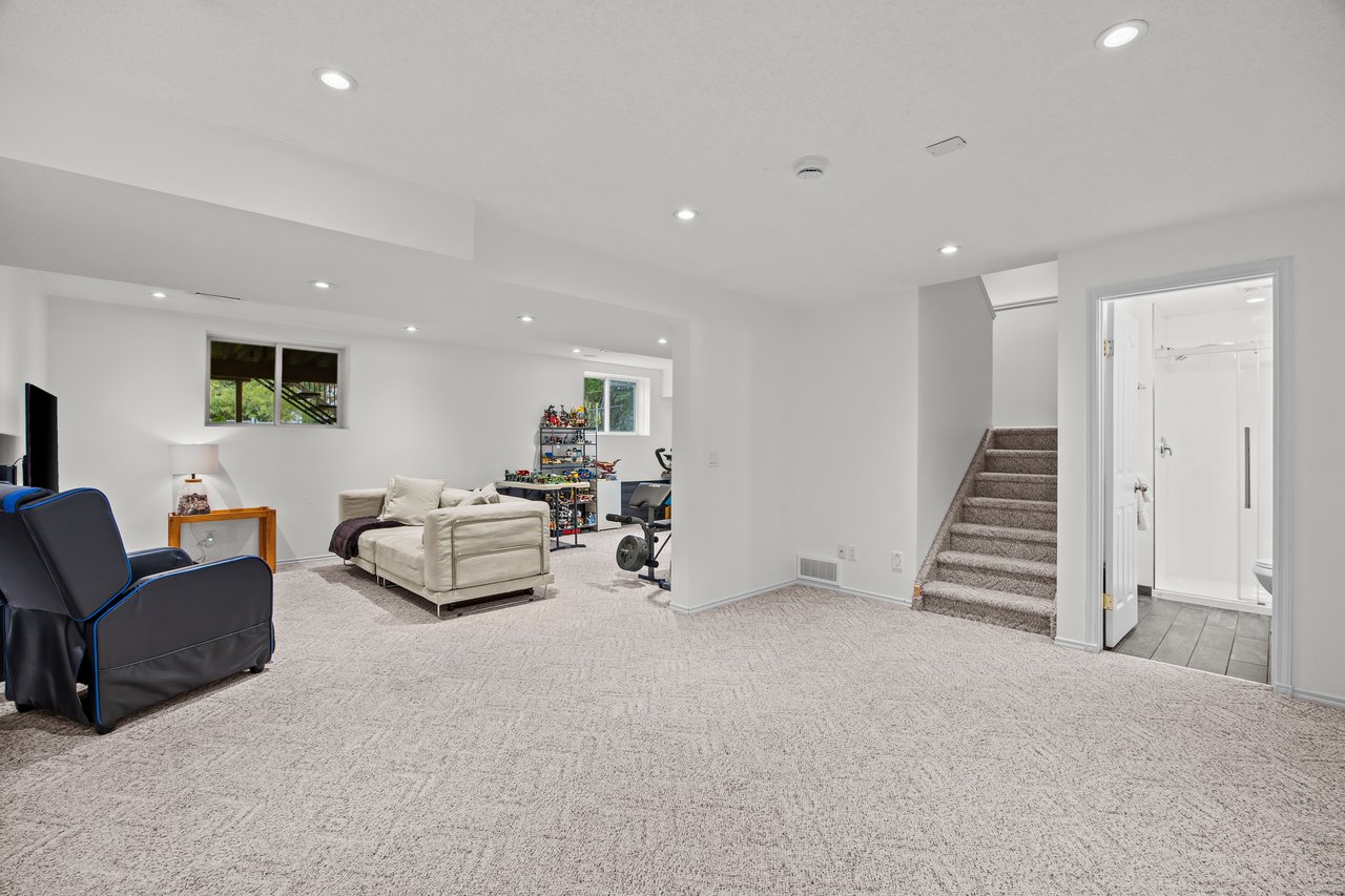 Basement recreation room with a cream sofa, recliner, toys, exercise equipment, stairs, and bathroom doorway.