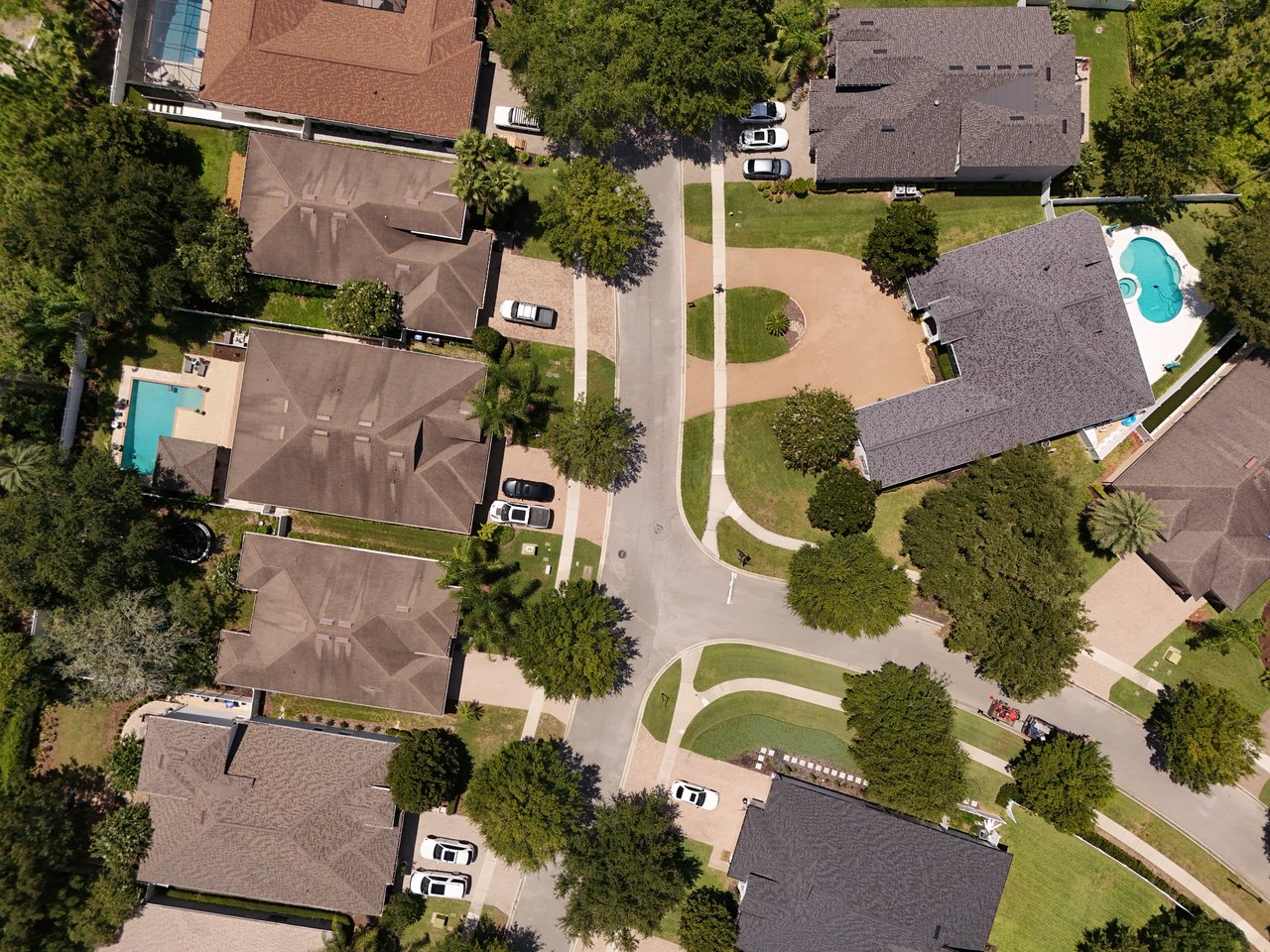 Aerial view of homes along a tree-lined residential street, with swimming pools in several backyards.