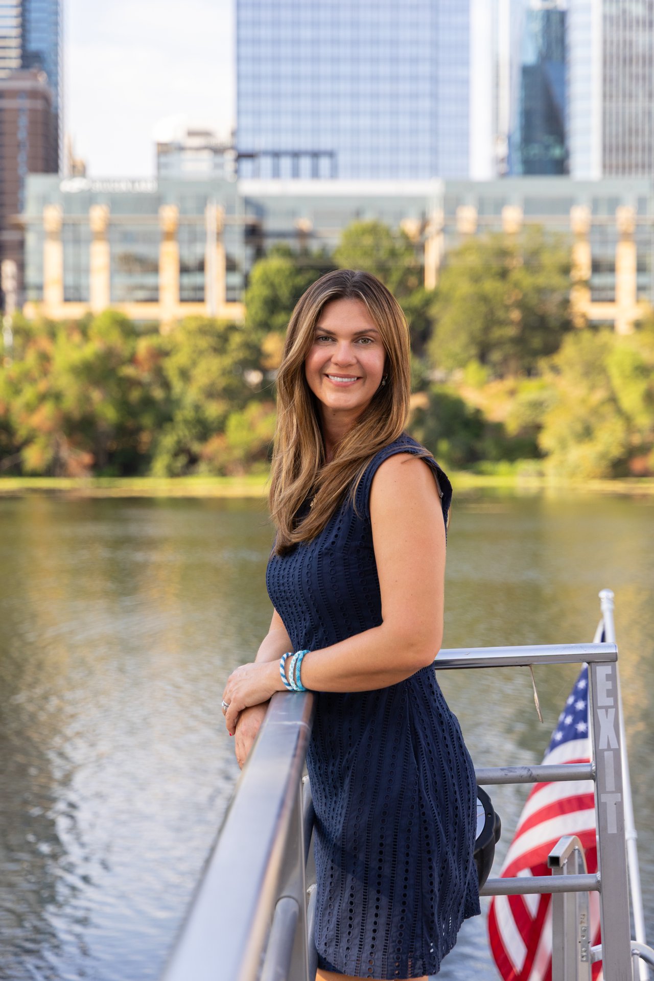 Woman in a navy dress standing beside a boat railing, with water, an American flag, and city buildings behind her.